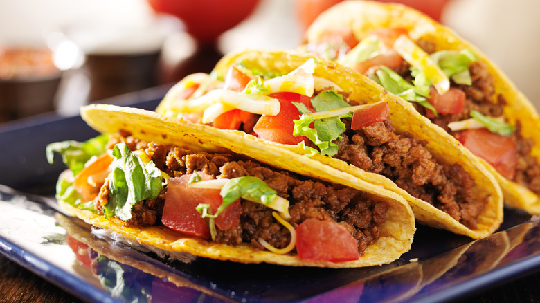 Three hard shell tacos filled with meat, tomatoes, lettuce, and cheese on a blue plate