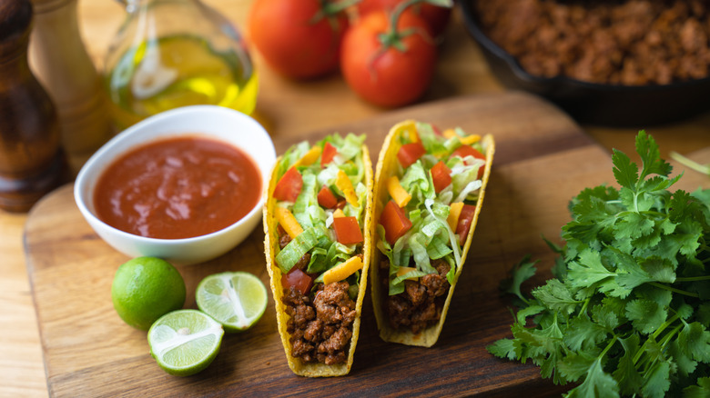 Two hard shells upright with meat, lettuce, tomato, and cheese surrounded by taco ingredients.