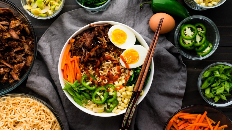 A colorful bowl of ramen with hardboiled eggs, carrots, jalepenos, shredded pork, and sauce, surrounded by bowls of separate ingredients