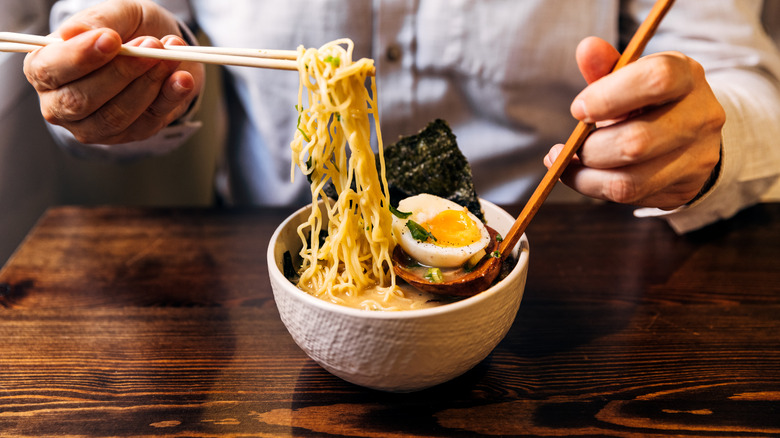 Chef's hands placing ramen, hardboiled egg, and seaweed in a white bowl