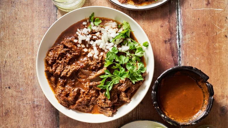 Birria in bowl with cilantro and onions surrounded by sides