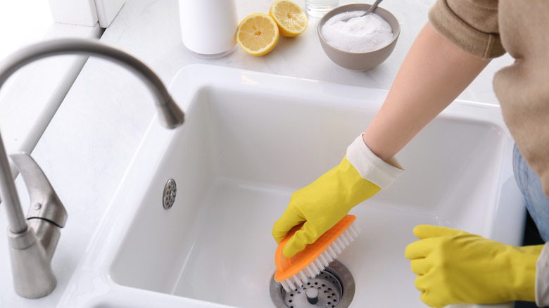 Someone wearing yellow rubber gloves and scrubbing their kitchen sink with baking soda and lemons nearby