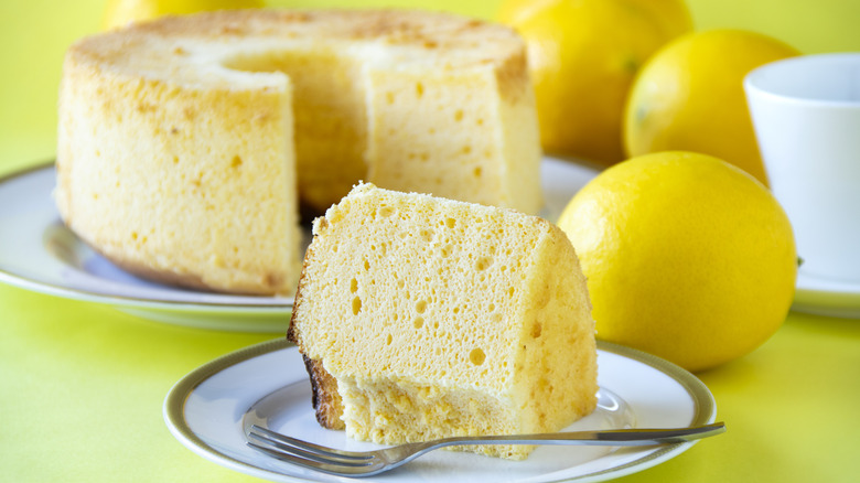 Slice of lemon chiffon cake in front of whole cake and lemons on lime background