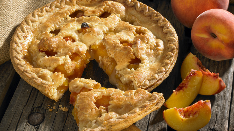 Slice cut out of peach pie next to peaches on wooden table