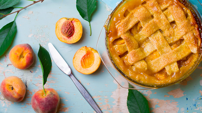 A whole latticework peach pie next to fresh peaches and leaves on a blue wooden table