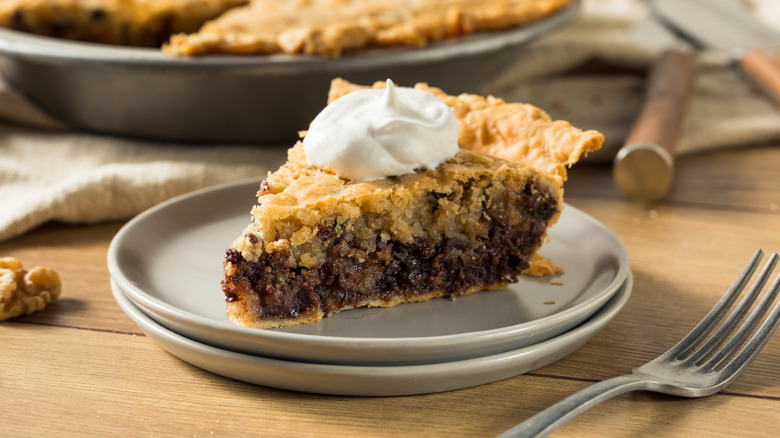 Two stacked beige plates with a slice of derby pie on wooden table with a fork on the side and a whole pie in the back