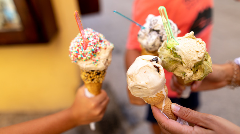 Hands holding four ice cream cones packed with different ice cream varieties with garnishes and plastic spoons