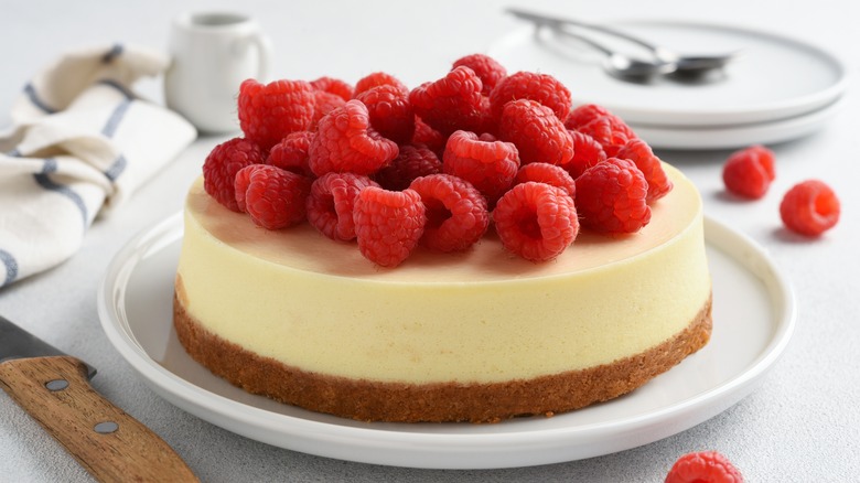 New York cheesecake topped with raspberries on white plate sitting on a white counter next to a knife with plates in the back