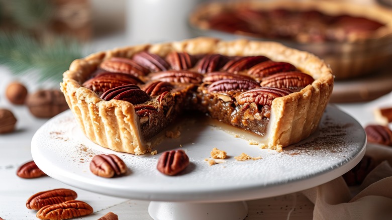 White cake stand holding a pecan pie with one slice removed surrounded by pecans and a pie in the back