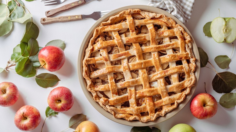 Apple pie with a lattice top crust surrounded by red apples on a white table