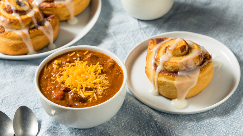 Bowl of chili with cheese next to cinnamon roll on light blue tablecloth