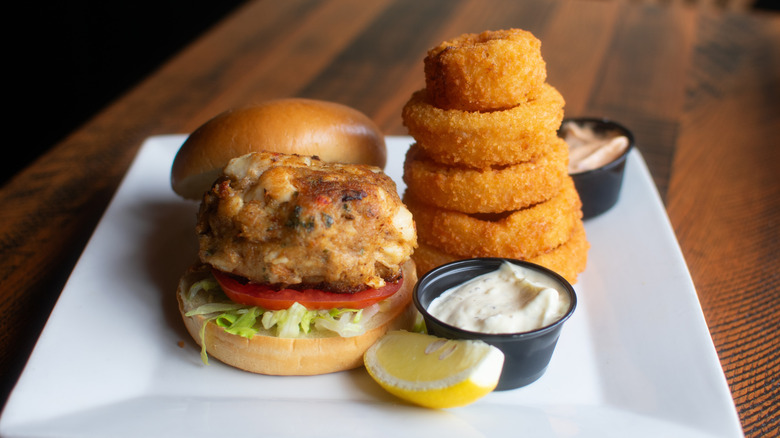 Crab cake sandwich with lettuce and tomato and side of onion rings on white plate