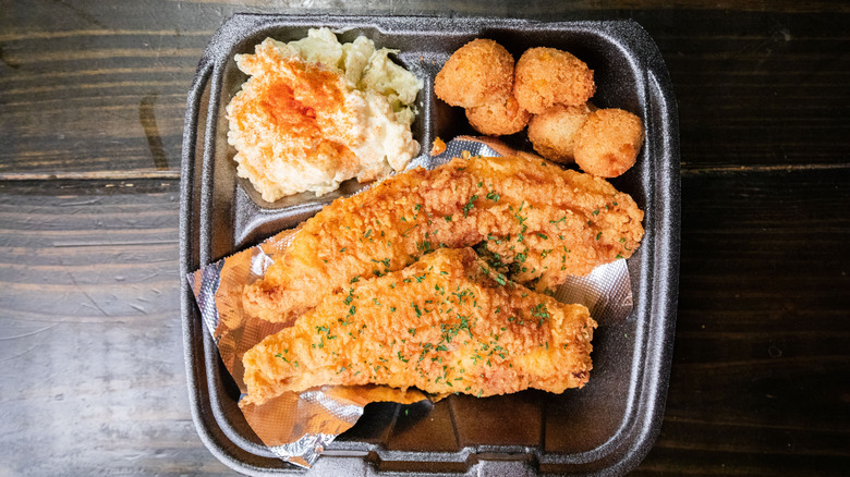 Fried catfish in black to-go container with mashed potatoes and hush puppies