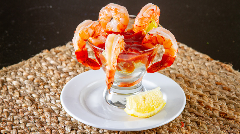 Shrimp hanging off cocktail glass full of sauce on white plate with lemon garnish