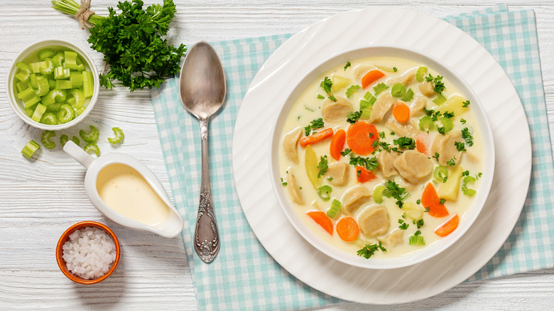 Creamy vegetable and dumpling soup on white table with diced veggies, salt, and herbs on the side