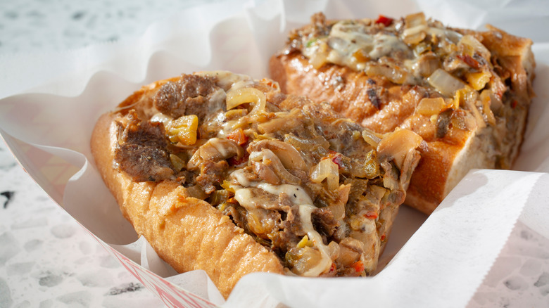 Halved Philly cheesesteak in paper container on white background