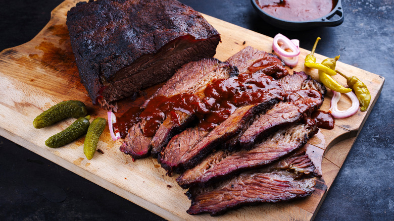 Sliced smoked brisket on cutting board with BBQ sauce, pickle, and pepper garnishes