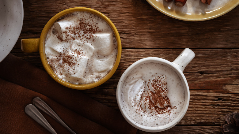 Two mugs of hot chocolate with marshmallows, cream, and cocoa powder on a wooden table