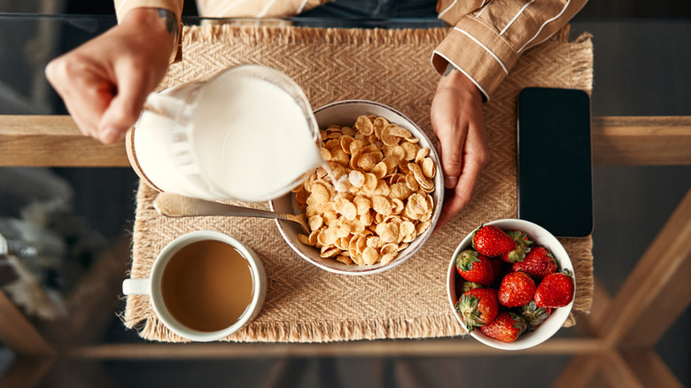 Overhead shot of hands pouring milk into bowl of cereal flakes, with a bowl of strawberries and a cup of coffee nearby