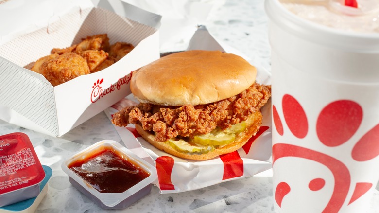 An open box of Chick-Fil-A nuggets, an open packet of barbeque sauce, a Chick-fil-A crispy chicken sandwich, and a Chick-Fil-A drink container laid out on a white table