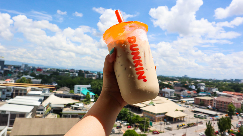 Hand holding a Dunkin' Donuts iced coffee with buildings and sky in the background.