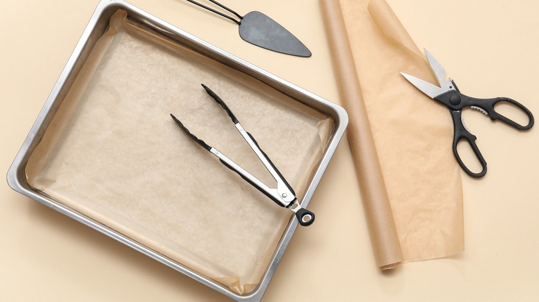 A baking tray with roll of parchment paper, tongs, and scissors