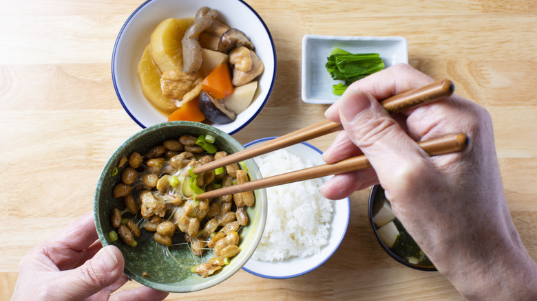 Hands holding a bowl of natto and chopsticks with other classic Japanese breakfast dishes in background