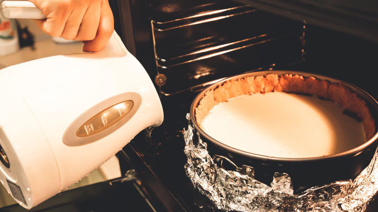 Hand holding a boiling pot pouring water into an oven's water bath with a cake tin inside