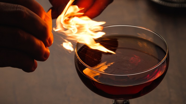 Hands holding a flaming orange peel over a Revolver cocktail