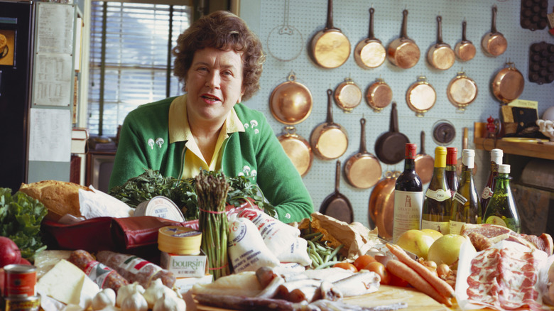 Julia Child behind kitchen counter piled with various meats, cheeses, and vegetables