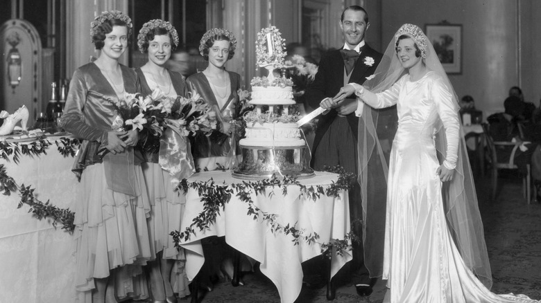 Bride and groom cutting into wedding cake alongside bridal party