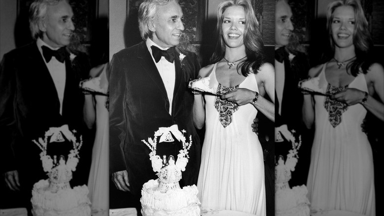 Bride holding piece of wedding cake while standing with groom beside cake in 1970s