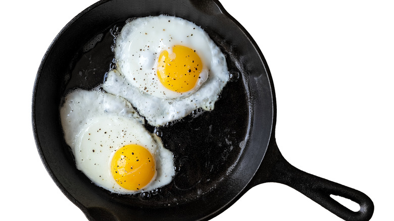 Two fried eggs with black pepper in a cast iron pan on white background