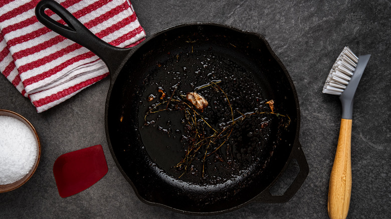 Dirty cast iron skillet being prepared for cleaning with coarse salt, brush, scraper and dish towel on a counter.