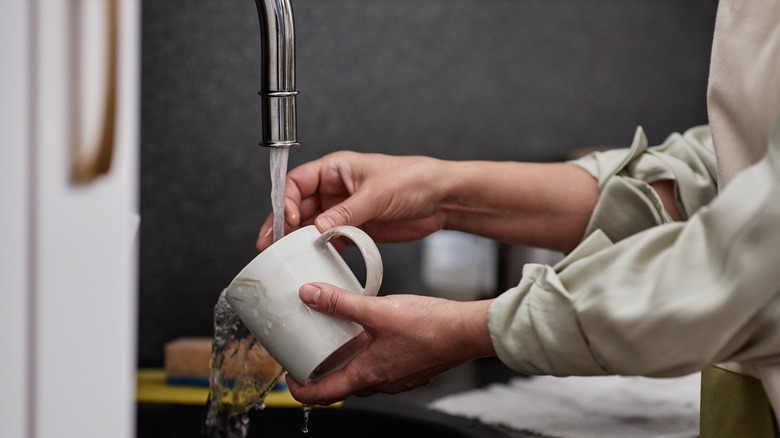 A woman washes a white coffee mug at the sink.