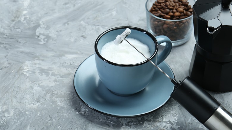 A handheld milk frother rests against a blue mug with a container of coffee beans behind it.