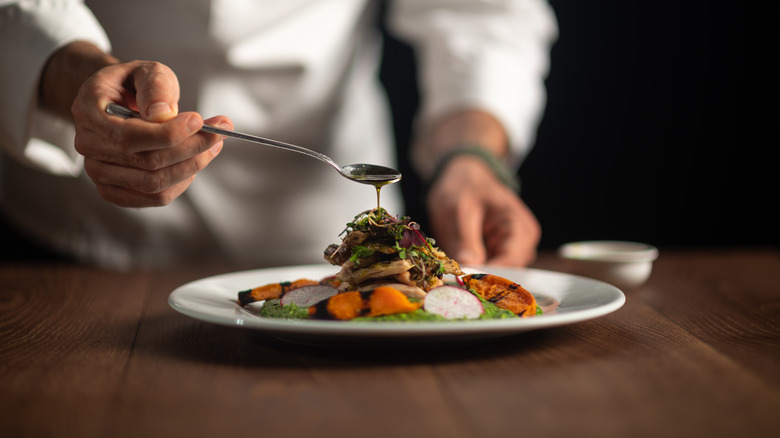 A chef using the spoon to pour a sauce on a dish that is assembled on a white plate