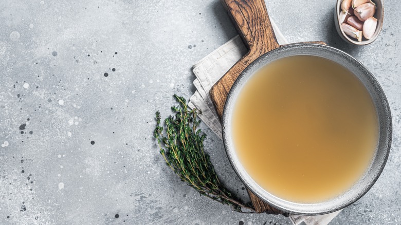 High angle view of a pan filled with homemade bone broth shot on rustic wooden board and a grey counter next to thyme and garlic