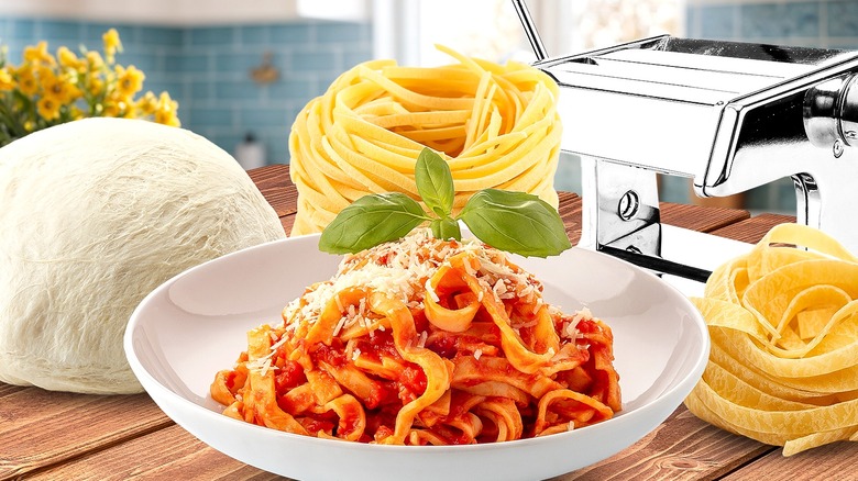 Pasta in different forms on a wooden table with a pasta machine in the background and blue kitchen backsplash