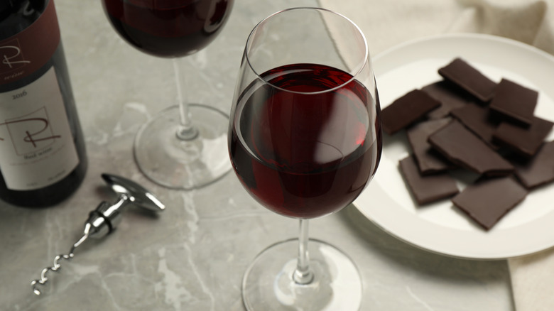 a glass of red wine in the front with a bottle of red wine, another glass of red wine, a plate with dark chocolate chunks, and a bottle opener in the back