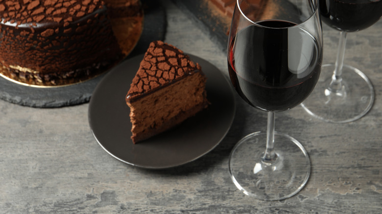 a piece of double chocolate cake on a plate placed next to a glass of red wine with the whole cake and in the background