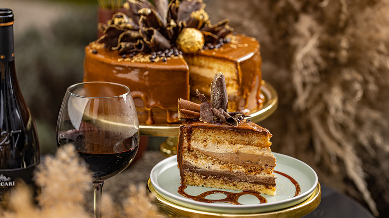 elaborate layered chocolate cake on a plate next to a glass of red wine and a wine bottle with the whole cake in the back