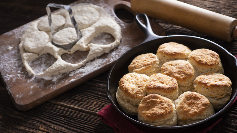 Buttermilk biscuits in a cast iron pan with a wooden cutting board with fresh biscuit dough on it and a rolling pin