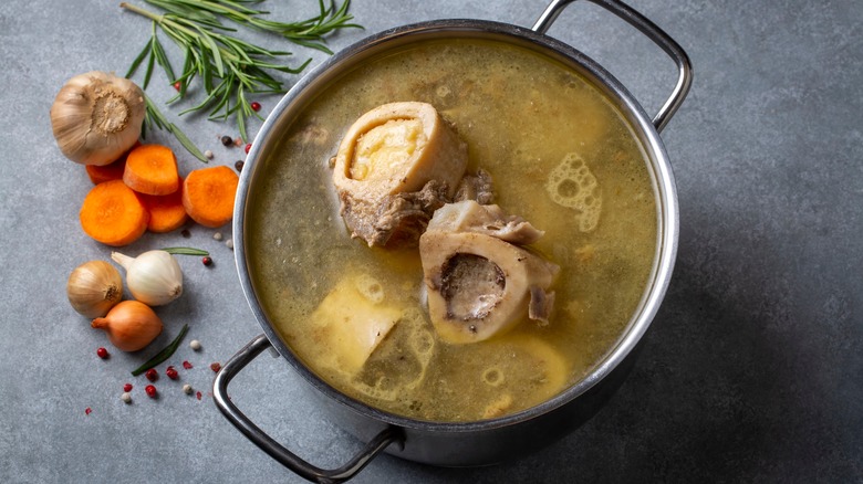 Pot of bone broth with marrow bones and a gray background with rosemary, garlic, and carrots