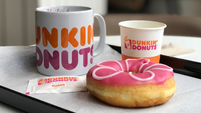 A Dunkin' branded mug, paper cup, pink donut, and sugar packet