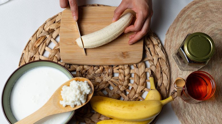 A bowl of kefir, a wooden spoon with kefir grains on it, and a hand slicing a fresh banana