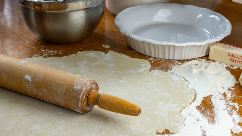 Wooden rolling pin, pie dough, empty pie tin, stick of butter, and flour on wood counter