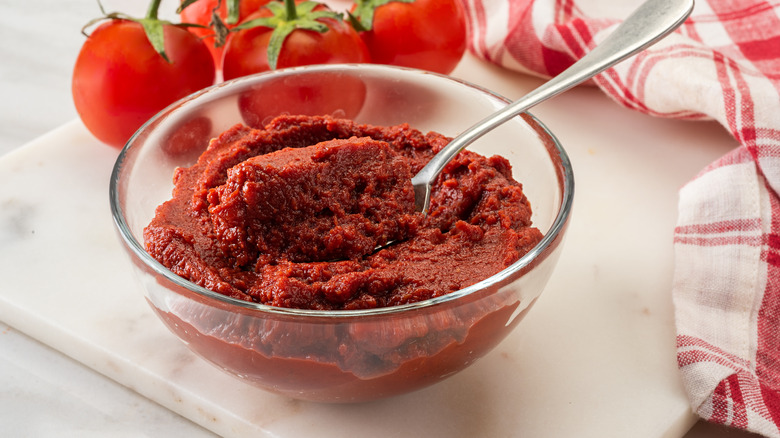 Thick tomato paste in a glass bowl with a spoon and fresh tomatoes in the background