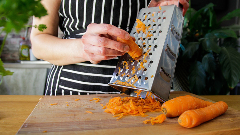 A person standing next to the kitchen counter grating fresh carrots on a wooden cutting board