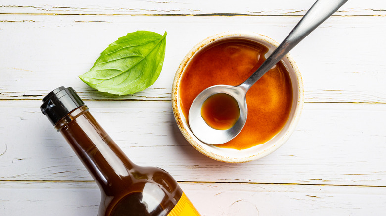 Bottle of Worcestershire sauce with some decanted into small dish and a green leaf all lined on white wood